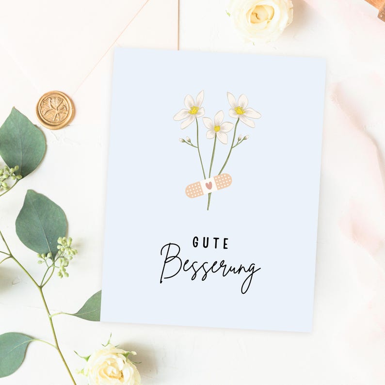 May include: A light blue card with a white flower bouquet and a bandage with a heart on it. The card says "Gute Besserung" which is German for "Get well soon".