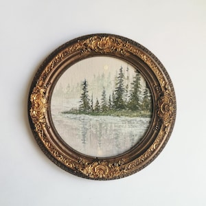 May include: Oval framed landscape painting featuring a serene forest scene reflected in calm water. The artwork is enclosed in an ornate, gold-toned frame with detailed floral and scrollwork embellishments. The painting's color palette includes muted greens, grays, and browns.