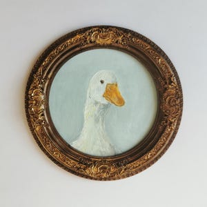 Könnte beinhalten: Gerahmtes Gemälde einer weißen Ente mit orangefarbenem Schnabel und hellblauem Hintergrund. Das Gemälde befindet sich in einem runden, verzierten, goldfarbenen Rahmen. Das Kunstwerk ist ein Porträt der Ente.