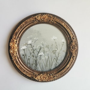May include: Oval framed artwork featuring a field of white wildflowers. The painting has a soft, muted color palette with green and gray tones. The ornate, gold-colored frame adds a vintage aesthetic to the piece.