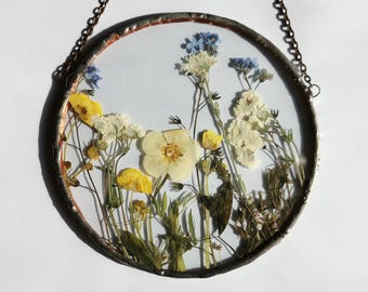May include: A round, clear glass piece with pressed wildflowers, including yellow, white, and blue blooms. The glass is framed with a metal edge and suspended by a chain, creating a decorative wall hanging.