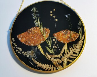 May include: A round, black wall hanging with a gold-colored frame and chain. The artwork features two orange and white spotted mushrooms surrounded by dried plants and flowers. The mushrooms are the focal point, set against a dark background.