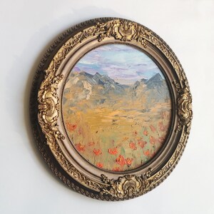 May include: A circular oil painting in a gold ornate frame. The landscape shows mountains, a field of red flowers, and a blue and purple sky. The frame has detailed carvings and a cream-coloured inner border.