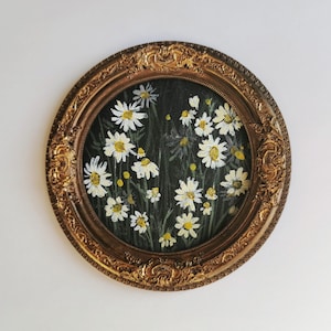 Puede incluir: Una pintura ovalada de margaritas blancas con centros amarillos sobre un fondo oscuro, enmarcada en un marco dorado ornamentado. La obra evoca una estética vintage, adecuada para la decoración del hogar.