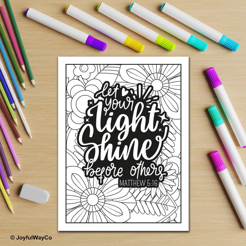 May include: A black and white floral coloring page with the text "let your light shine before others" and the reference "Matthew 5:16". Colored pencils and markers in various colors are arranged around the page on a wooden surface.