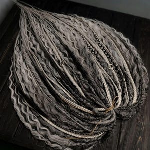Pode incluir: Um conjunto de dreadlocks sintéticas em preto, cinza e branco. As dreadlocks são onduladas e têm uma aparência texturizada.