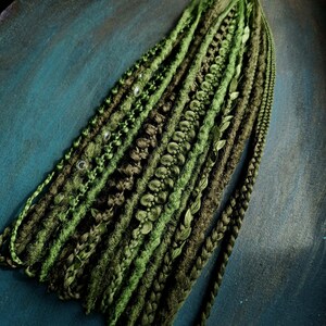 May include: A collection of synthetic dreadlocks in various shades of green and olive. The set includes textured twists, braided strands, and smooth locks, some accented with small silver beads and delicate leaf-like embellishments throughout.