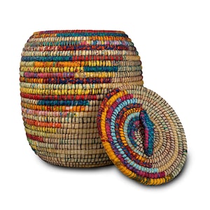 Puede incluir: Una cesta redonda tejida con tapa. La cesta est&aacute; hecha de fibras naturales y presenta un dise&ntilde;o de patchwork colorido con tonos de rojo, amarillo, azul y verde. La tapa es circular y est&aacute; al lado de la cesta.