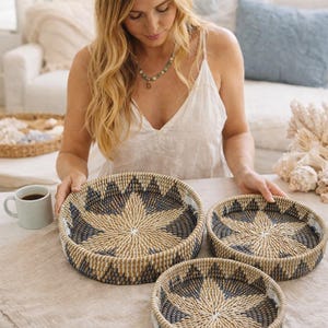May include: Three round woven baskets with a starburst design in natural, tan, and navy blue. The baskets are on a light-colored surface. The baskets are of different sizes and appear to be handmade.
