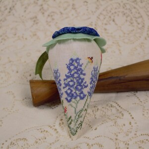 May include: A white fabric pincushion shaped like a cone with blue and green fabric accents. The pincushion is embroidered with blue flowers and green leaves. It is sitting on a white lace tablecloth.