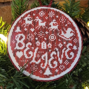 Peut inclure: Une décoration de Noël ronde en point de croix rouge et blanc. L'ornement présente le mot "BELIEVE" en blanc, ainsi que des rennes, des flocons de neige et d'autres motifs sur le thème des fêtes. Une ficelle rouge est attachée pour l'accrocher.