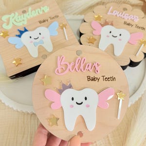 May include: Three wooden baby tooth keepsake boxes. Each box features a smiling tooth with wings, a crown, and a magic wand. The boxes are personalized with names in pastel colors and the words "Baby Teeth".