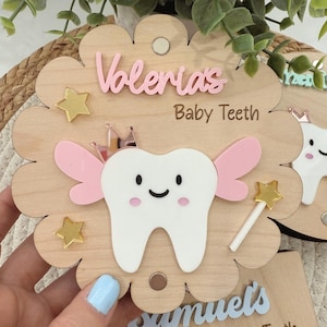 May include: A wooden tooth chart with a pink tooth fairy design. The chart has a pink tooth with a crown, pink wings, and a gold star wand. The chart also has a gold star and the text "Valerias Baby Teeth".