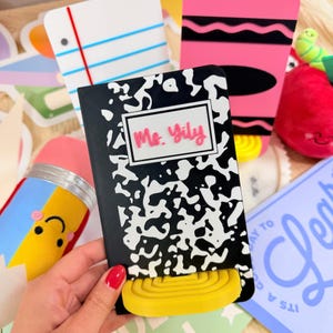 Puede incluir: Un letrero de acrílico en blanco y negro estilo cuaderno de composición con una etiqueta rectangular blanca con el texto Ms. Yily en letra cursiva rosa. El letrero tiene una base amarilla semicircular acanalada y otros adornos escolares.