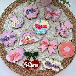 Puede incluir: Una colección de 12 adornos de madera personalizados con diversas formas y colores. Los adornos presentan nombres como Mia, Valeria, Mylana, Reese e Isabella. Cada adorno tiene un diseño único, incluyendo una flor, un corazón, un arcoíris, una concha, una mariposa, una cereza y una bailarina.