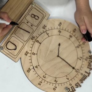 K&ouml;nnte beinhalten: Eine Holzuhr mit einem runden Zifferblatt und einem beweglichen Stundenzeiger. Das Zifferblatt der Uhr hat Zahlen von 1 bis 12 und Markierungen f&uuml;r jeweils 5 Minuten. Die Uhr hat auch einen separaten Abschnitt mit drei Schlitzen zum Einstellen der Zeit, wobei die Zahlen 1, 0 und 8 sichtbar sind. Der Text "What Time Is It" ist auf der Uhr gedruckt.