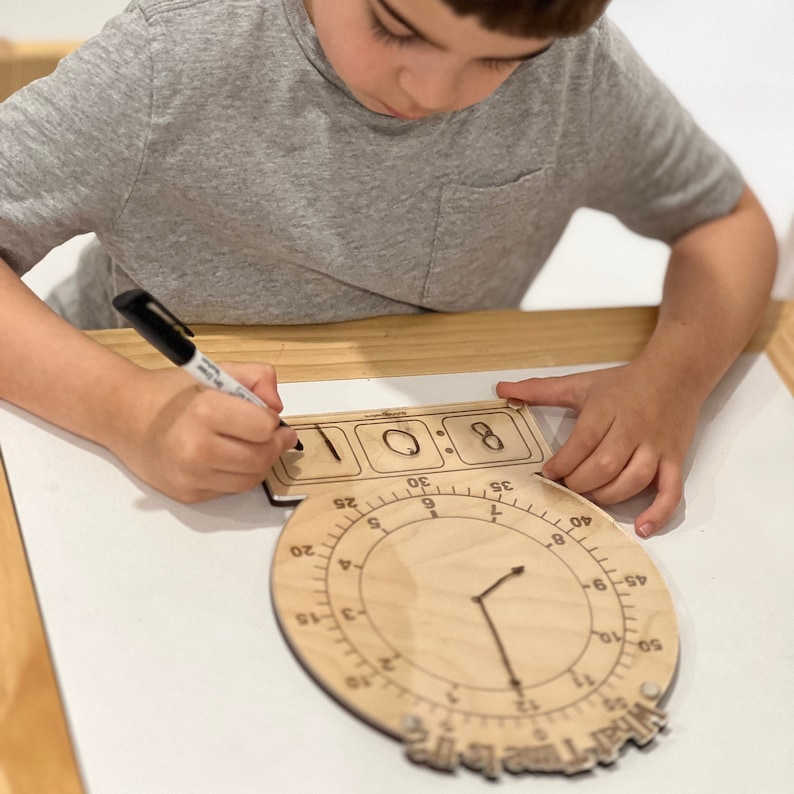 K&ouml;nnte beinhalten: Eine Holzuhr mit einem Zifferblatt und einer digitalen Zeitanzeige. Das Zifferblatt der Uhr hat Zahlen von 1 bis 12 und Markierungen f&uuml;r Minuten. Die digitale Zeitanzeige zeigt 8:00. Ein Kind schreibt mit einem schwarzen Stift auf die digitale Zeitanzeige.