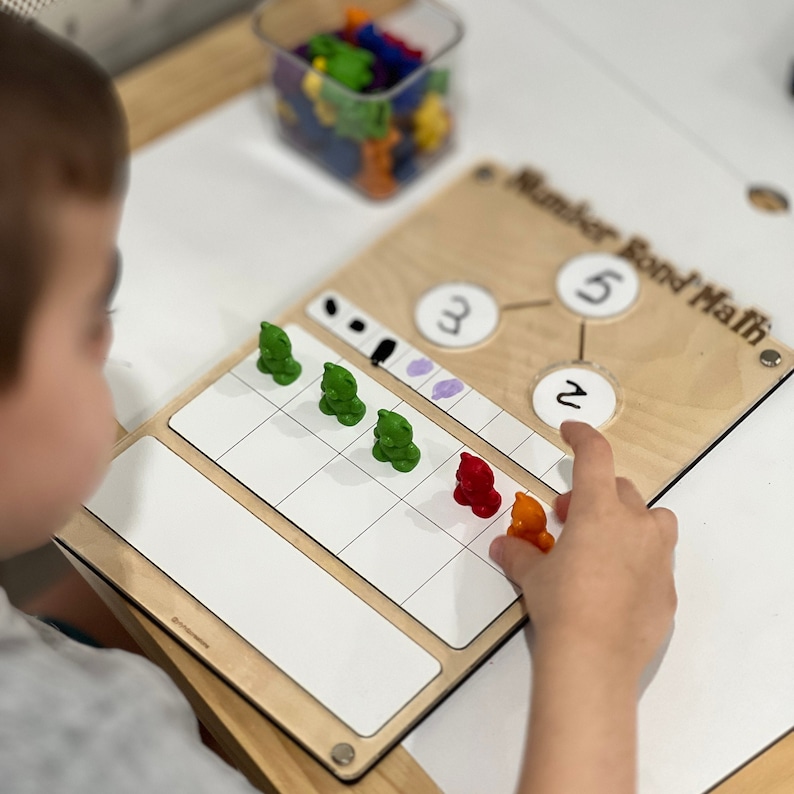 Pu&ograve; includere: Un bambino sta imparando le unioni numeriche con un pannello di legno su cui &egrave; scritto "Number Bond Math". Il pannello ha tre cerchi con i numeri 3, 5 e 2 scritti al loro interno. Il bambino sta usando gettoni colorati per rappresentare i numeri.