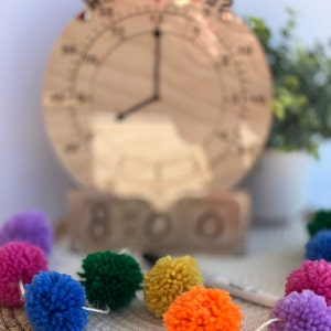 K&ouml;nnte beinhalten: Eine Holzuhr mit einer Regenbogen-Pompon-Girlande. Das Zifferblatt der Uhr ist verschwommen und die Girlande ist scharf. Das Zifferblatt der Uhr hat die Zahlen 1-12 und das Wort "BOO" ist auf dem Sockel der Uhr geschrieben.