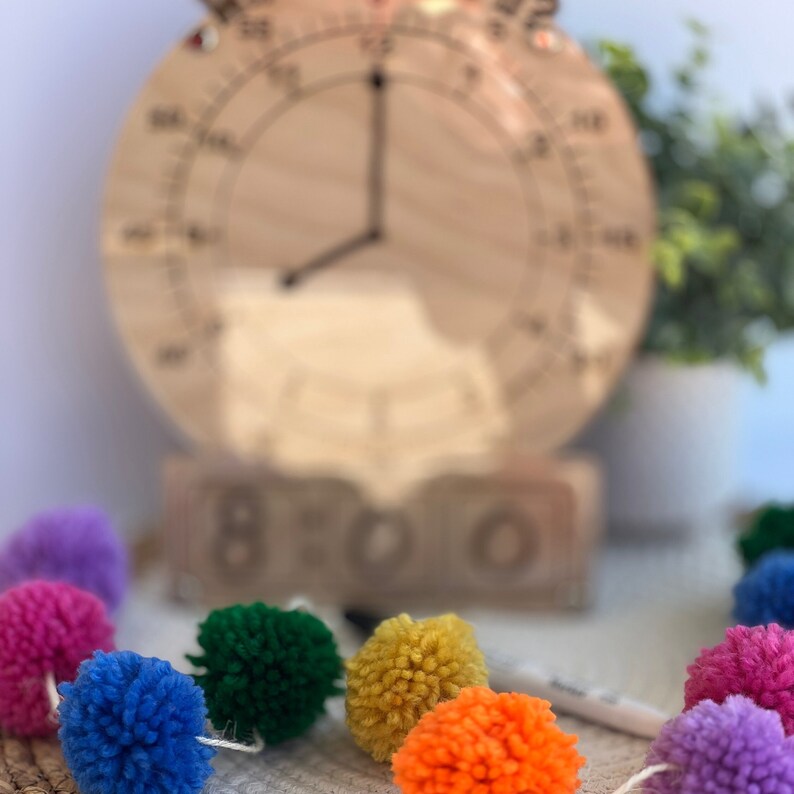 K&ouml;nnte beinhalten: Eine Holzuhr mit einer Regenbogen-Pompon-Girlande. Das Zifferblatt der Uhr ist verschwommen und die Girlande ist scharf. Das Zifferblatt der Uhr hat die Zahlen 1-12 und das Wort "BOO" ist auf dem Sockel der Uhr geschrieben.