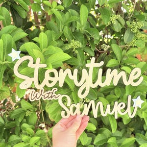 Puede incluir: Letrero de madera clara con las palabras "Storytime with Samuel" en fuente cursiva. El letrero tiene detalles de estrellas y está sobre un fondo de follaje verde.