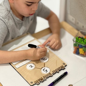 Pu&ograve; includere: Un bambino usa un pennarello cancellabile a secco per scrivere su una lavagna di matematica a legami numerici in legno. La lavagna ha tre cerchi con i numeri 2, 3 e 5 scritti all'interno. La lavagna ha anche una griglia con caselle vuote. Il bambino &egrave; seduto a un tavolo con un contenitore di manipolatori di conteggio colorati.