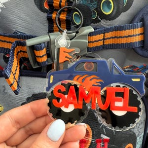 Może przedstawiać: Niebieski brelok do kluczy w kształcie monster trucka z imieniem "SAMUEL" w czerwonych literach. Ciężarówka ma pomarańczowe płomienie i czarno-białe koła. Brelok jest przymocowany do paska plecaka z pomarańczowymi i niebieskimi akcentami.