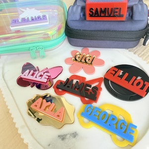 May include: A collection of personalized acrylic name tags in various shapes and colors, including a butterfly with Alice, a flower with Gigi, a truck with James, a circle with Elliot, a cloud with Ayla, and a car with George, plus a Samuel tag.