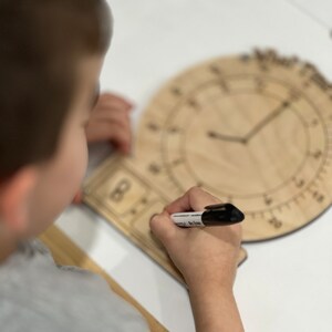 K&ouml;nnte beinhalten: Ein Kind benutzt einen schwarzen Trockenl&ouml;schmarker, um auf ein h&ouml;lzernes Zifferblatt zu schreiben. Das Zifferblatt hat einen gro&szlig;en Kreis mit Zahlen am Rand und einen kleineren Kreis in der Mitte mit einem Zeiger, der auf die Zahl 8 zeigt.