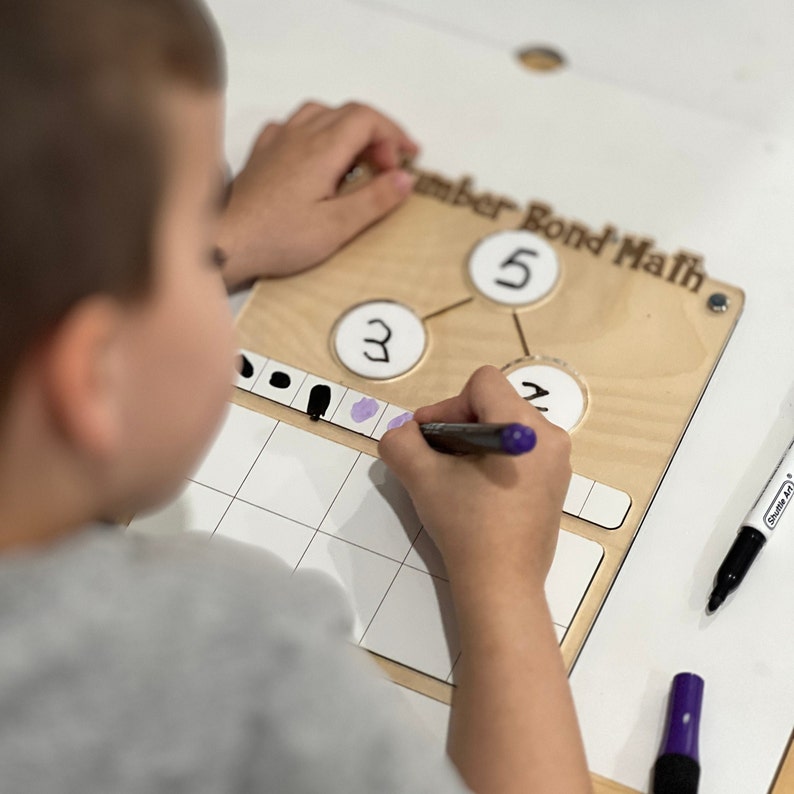 Pu&ograve; includere: Un bambino usa un pennarello viola per completare un'attivit&agrave; di matematica di legame numerico su una lavagna di legno. La lavagna ha tre cerchi con i numeri 3, 2 e 5 scritti al loro interno. Il bambino sta scrivendo punti su una griglia per rappresentare il numero 5.