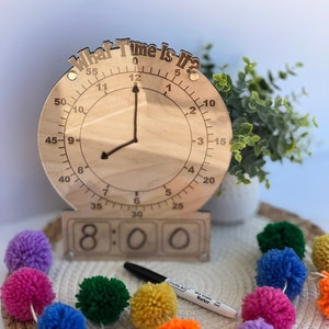 K&ouml;nnte beinhalten: Eine Holzuhr mit einem beweglichen Zeiger und einer separaten Anzeige f&uuml;r Stunden und Minuten. Das Zifferblatt der Uhr lautet "What Time Is It?" und die Anzeige zeigt 8:00. Die Uhr ist von bunten Pompons umgeben.
