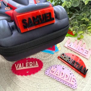 Könnte beinhalten: Eine Sammlung personalisierter Namensschilder in verschiedenen Formen und Farben. Die Schilder enthalten Namen wie "SAMUEL", "VALERIA", "ALICE", "CHRISTIAN" und "MADDIE". Die Schilder sind rot, rosa und lila mit schwarzer Schrift. Im Hintergrund befindet sich ein schwarzes Federmäppchen.