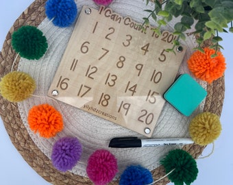 May include: A wooden learning board with the text "I Can Count To 20" and numbers 1 through 20 written in black. The board is surrounded by a colorful garland of pom-poms. A dry erase marker and a small green eraser are also included.