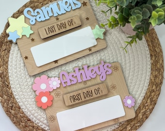 May include: Two wooden signs with blank spaces for writing. The top sign is light brown with a blue name "Samuels" and the text "Last Day Of:". The bottom sign is light brown with a pink name "Ashley's" and the text "First Day Of:". Both signs have decorative stars and flowers.
