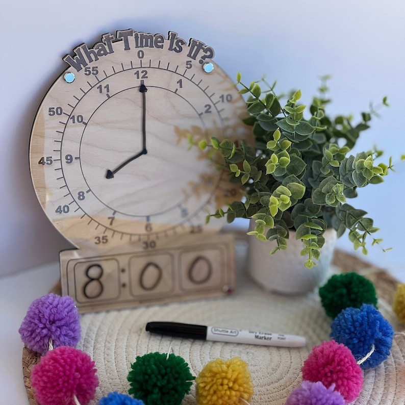 K&ouml;nnte beinhalten: Eine Holzuhr mit beweglichen Stunden- und Minutenzeigern, eine Flip-Clock, die 8:00 anzeigt, und ein blauer und wei&szlig;er Stift. Die Uhr tr&auml;gt die Aufschrift "What Time Is It?" Um die Uhr und den Stift herum liegen bunte Pompons.