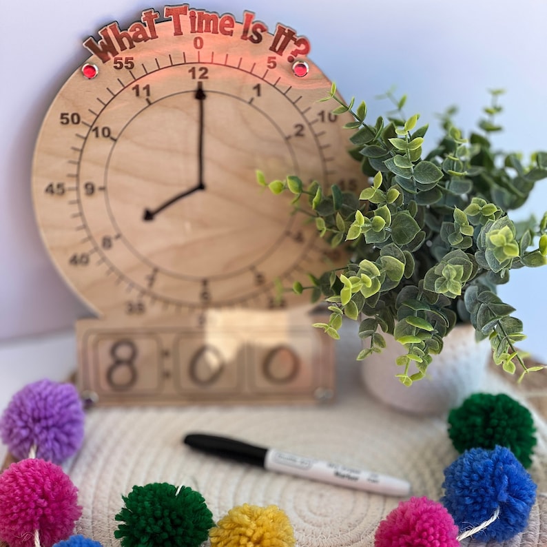 K&ouml;nnte beinhalten: Eine Holzuhr mit einem roten Schild "What Time Is It?", einem Satz Holznummernbl&ouml;cken, die 8:00 anzeigen, und einem schwarzen Stift auf einer wei&szlig;en Oberfl&auml;che mit bunten Pompons.
