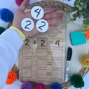 Puede incluir: Una herramienta de aprendizaje de madera con una cubierta de acr&iacute;lico transparente. La herramienta presenta un problema de matem&aacute;ticas de enlace num&eacute;rico con la ecuaci&oacute;n 2 + 2 = 4. Hay tres c&iacute;rculos blancos con los n&uacute;meros 2, 2 y 4 escritos en ellos. La herramienta est&aacute; sobre una superficie tejida con pompones de colores.