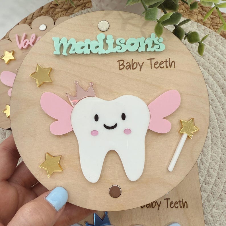 May include: A wooden tooth fairy box with a light wood background. The box has a white tooth with pink wings and a gold crown on top. The box also has gold stars and a wand with a gold star on top. The box says "Madison's Baby Teeth" on the front.