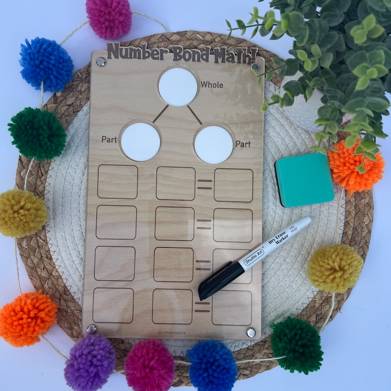 Puede incluir: Herramienta de aprendizaje de matem&aacute;ticas de enlace num&eacute;rico de madera con c&iacute;rculos y cuadrados para escribir n&uacute;meros. El tablero tiene las palabras "Number Bond Math!" en la parte superior y "Whole", "Part" y "Part" etiquetados en los c&iacute;rculos. Se incluye un marcador de borrado en seco.