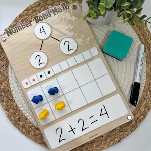 Könnte beinhalten: Holz-Zahlenbindungs-Mathetafel mit dem Text "Number Bond Math". Die Tafel hat ein Zahlenbindungsdiagramm mit den Zahlen 4 und 2 sowie die Gleichung 2+2=4. Enthält blaue und gelbe Zählsteine, einen grünen Radierer und einen Whiteboard-Marker.