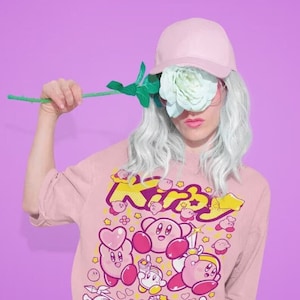 Peut inclure: Un sweat-shirt rose à manches longues avec un imprimé graphique de personnages Kirby. Le sweat-shirt est de couleur rose, jaune et blanche. Le texte "Kirby" est imprimé en gros caractères gras. Le modèle porte une casquette de baseball rose et a les cheveux blancs.