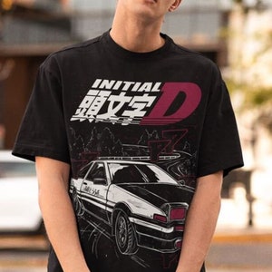 Puede incluir: Camiseta negra con un estampado gráfico blanco y rojo de un coche y el texto "INITIAL D" en japonés e inglés.
