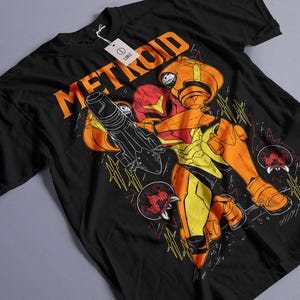Puede incluir: Camiseta negra con un gráfico de un personaje de la serie de videojuegos Metroid. El diseño incluye la palabra "METROID" en naranja y una ilustración detallada del personaje en naranja, amarillo y rojo.