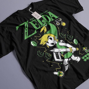 Puede incluir: Camiseta negra con un gráfico verde y amarillo de un personaje de videojuego con espada y escudo. El texto "THE END OF ZELDA" está impreso en verde. Una etiqueta blanca está adherida a la camiseta.
