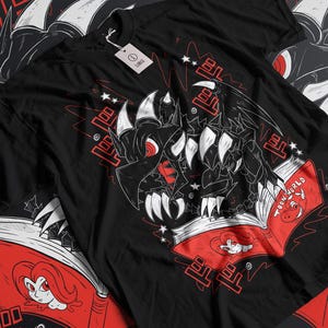 Op de afbeelding: Zwart T-shirt met een afbeelding van een zwarte draak die een rood boek vasthoudt met de tekst "Toon World" op de omslag. De draak heeft scherpe tanden en klauwen. De achtergrond is zwart met rode en witte sterren.