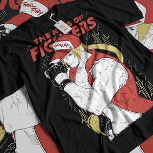 May include: Black t-shirt with a red and white illustration of a character from the video game "The King of Fighters". The character is wearing a red shirt and a red cap with the words "Fatal Fury" on it. The text "The King of Fighters" is written in white on the shirt.