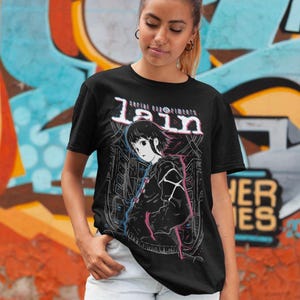 May include: Black t-shirt with a graphic print of a character from the anime series "Serial Experiments Lain". The graphic features the character Lain Iwakura in a black jacket with a pink and blue design. The text "Serial Experiments Lain" is printed above the character.