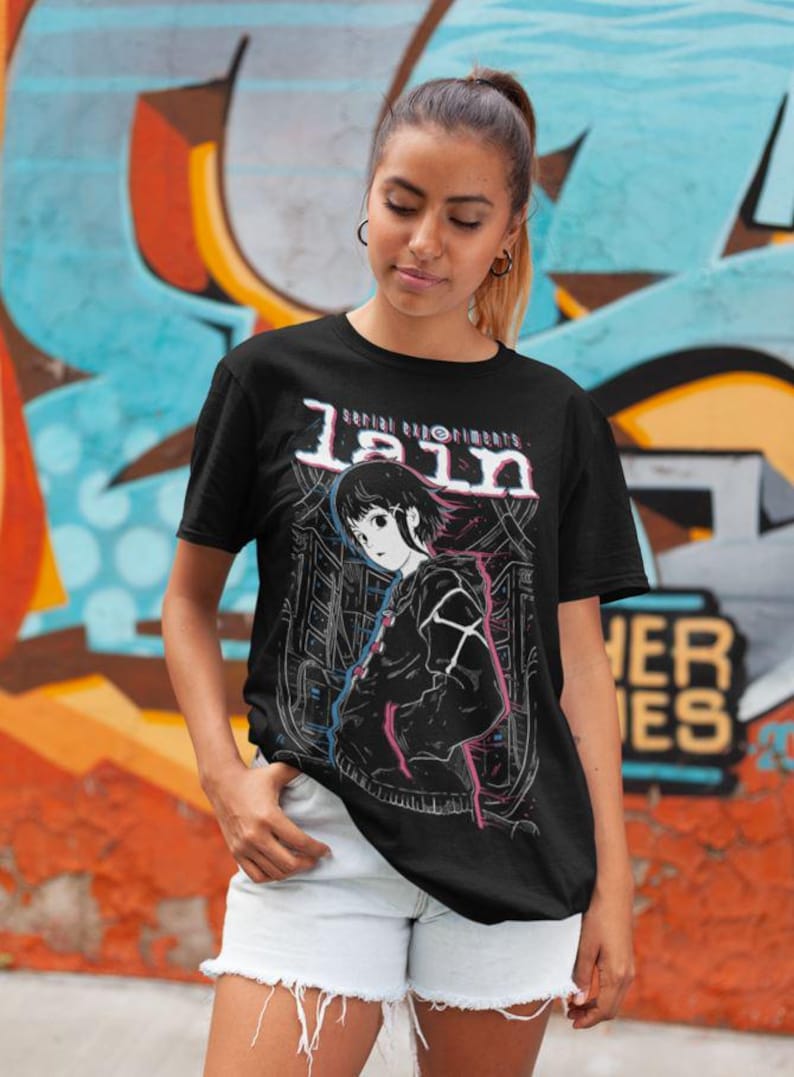 May include: Black t-shirt with a graphic print of a character from the anime series "Serial Experiments Lain". The graphic features the character Lain Iwakura in a black jacket with a pink and blue design. The text "Serial Experiments Lain" is printed above the character.