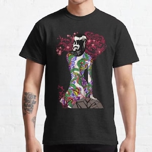May include: A black t-shirt with a colorful illustration of a woman with a tattoo of a snake and flowers. The woman is sitting on a rock with her back to the viewer.