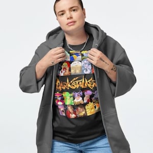 May include: A person wearing a dark gray zip-up hoodie over a black t-shirt with a colorful graphic print of various characters and the text "Darkstalkers". The person is also wearing blue jeans.